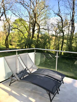 A glass-enclosed outdoor seating area with a metal frame, surrounded by a railing, overlooking a forested landscape. - Gîte "Fleur de Miel" – Renovated House in the Heart of the Lakes Region (Saffloz)