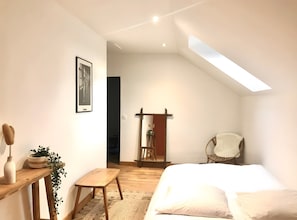 A bedroom with a bed, a wooden bench, a chair, a small table with a plant, and a framed picture on the wall. - Gîte "Fleur de Miel" – Renovated House in the Heart of the Lakes Region (Saffloz)