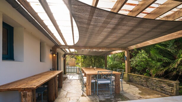Un patio al aire libre con una mesa y sillas de madera, rodeado de suelo de piedra y una pérgola de madera.