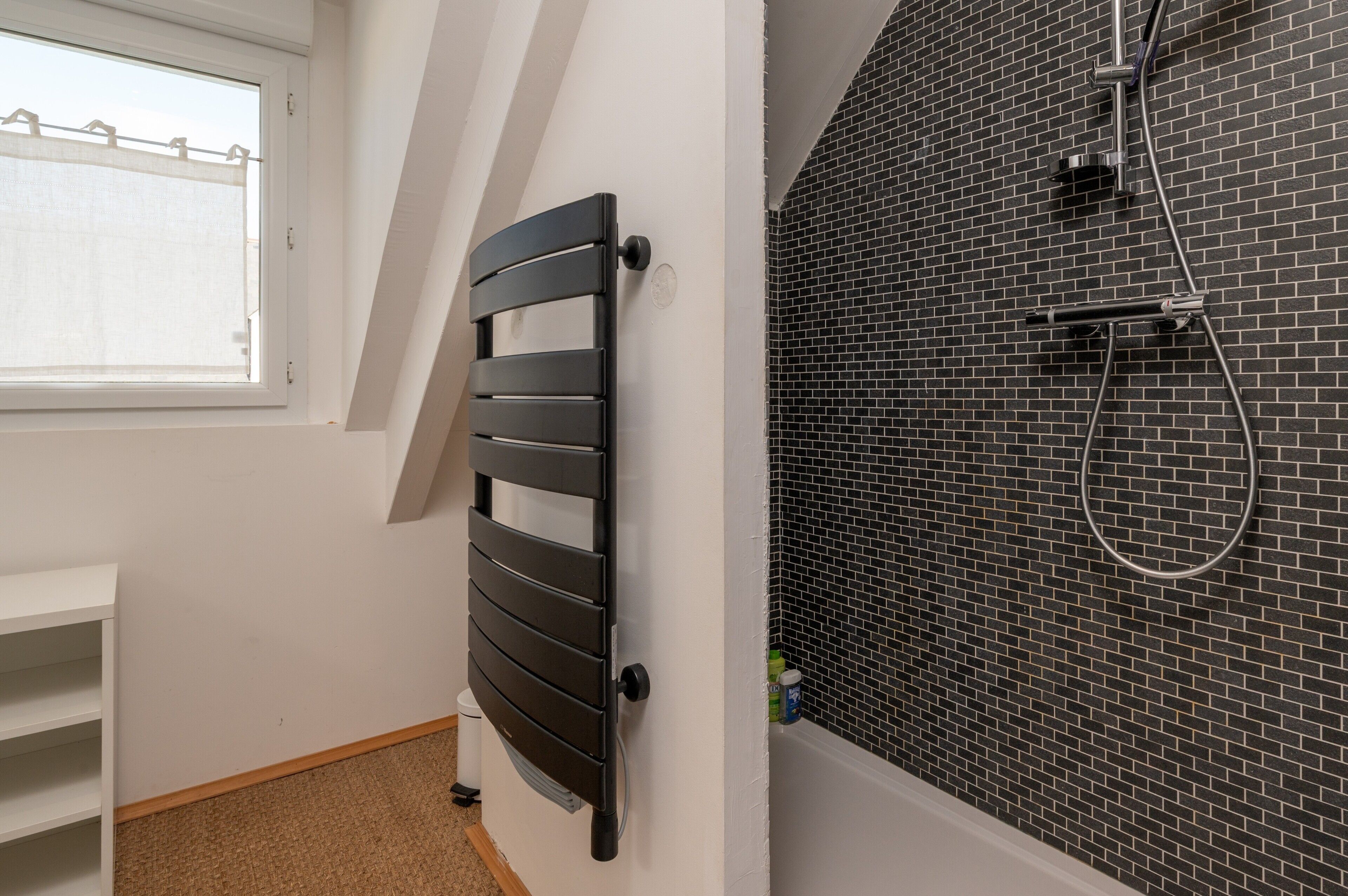 Une salle de bain avec un mur de douche recouvert de carreaux noirs, un radiateur noir et une fenêtre avec un store blanc.