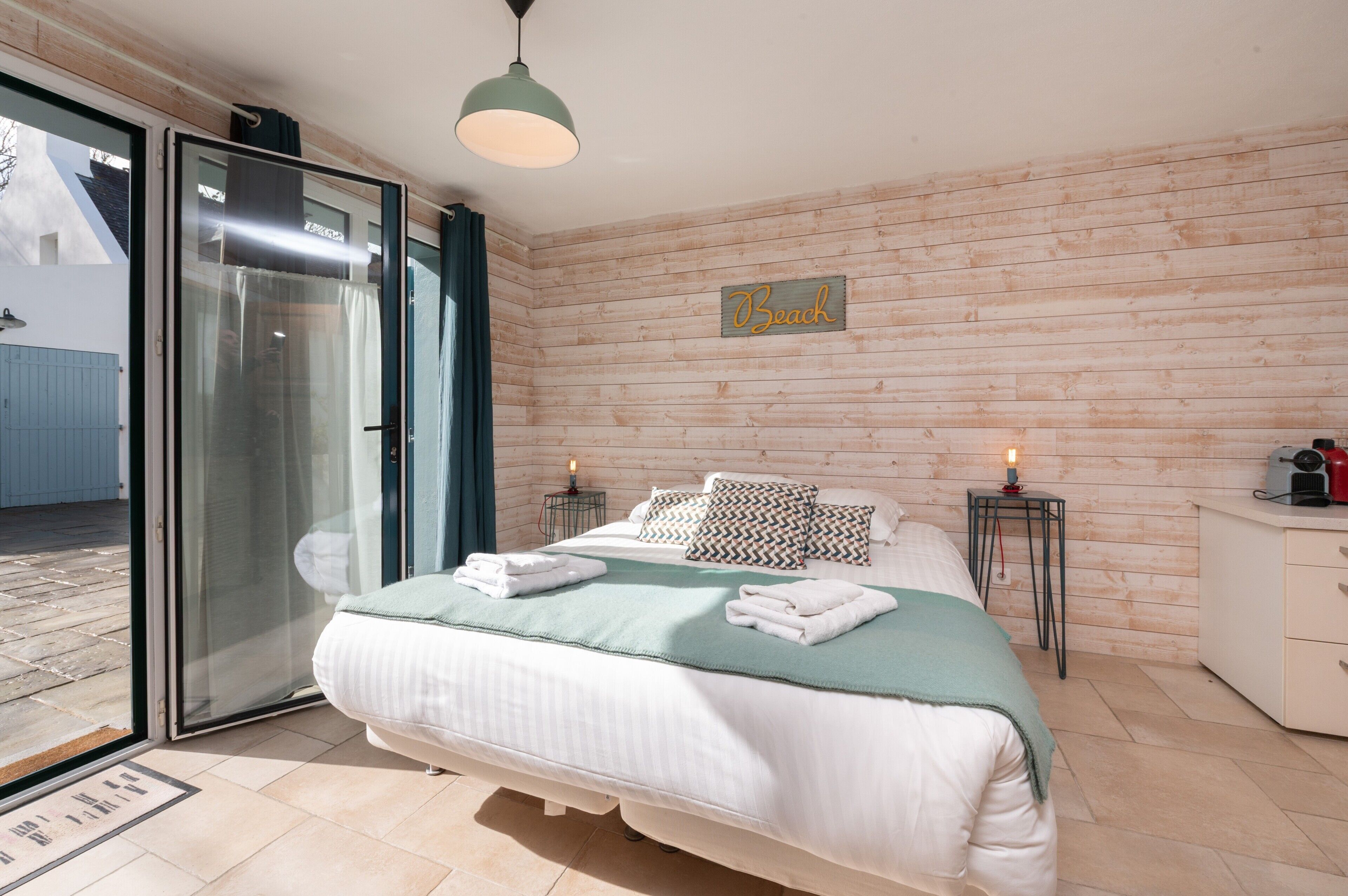 A bedroom with a bed, two bedside tables, a nightstand, a wooden wall, a sliding glass door, and a sign that says 'Beach'.
