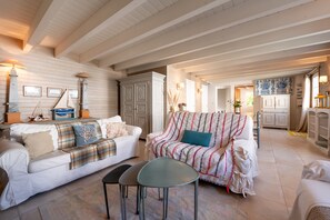 A cozy living room with a white sofa, a patterned armchair, and a small table. There is a wooden ceiling with exposed beams and a kitchen area in the background. - An Exceptional Property for Family or Friends’ Holidays on Belle-Île-en-Mer (Le Palais)