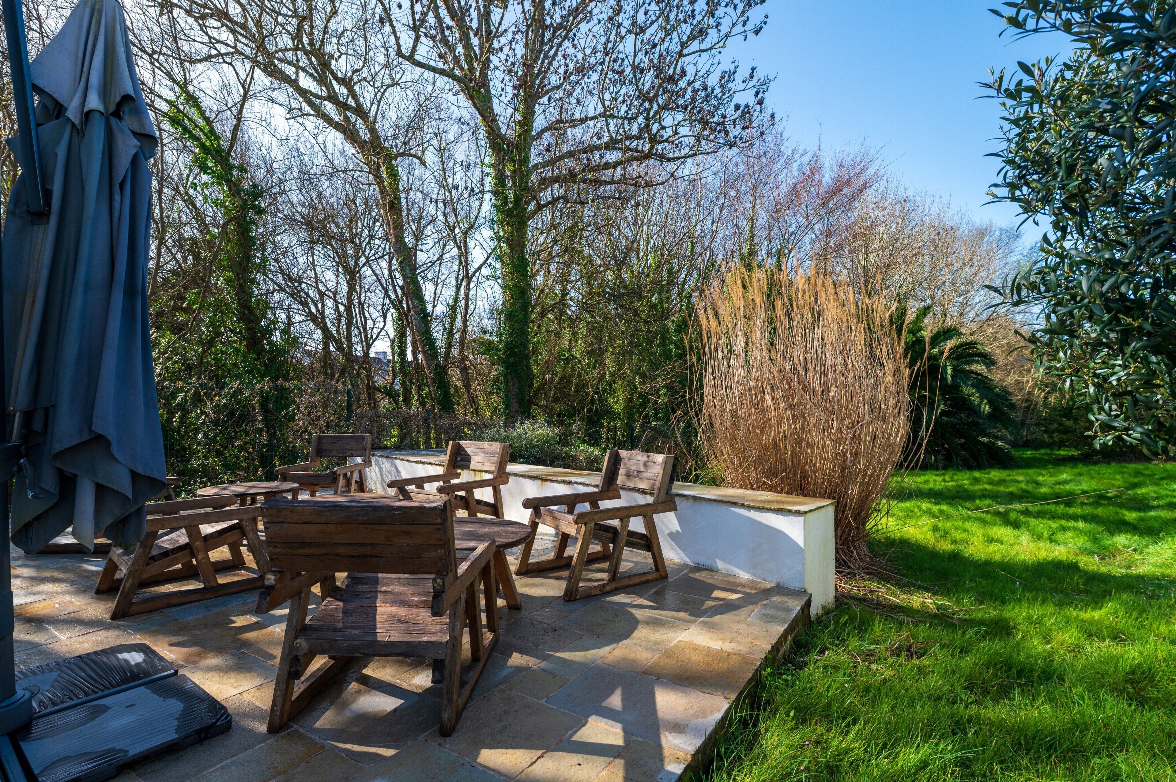 Une terrasse dotée de mobilier en bois, d’une table et de chaises, entourée d’un jardin avec des arbres et de la pelouse.
