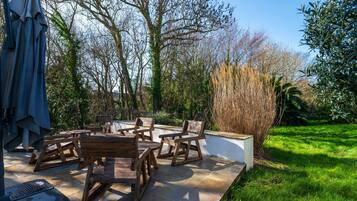 Une terrasse dotée de mobilier en bois, d’une table et de chaises, entourée d’un jardin avec des arbres et de la pelouse.