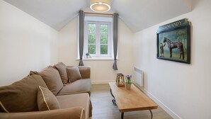 Ett rum med en brun soffa, ett träbord, ett fönster med gardiner, en taklampa och en inramad tavla på väggen.