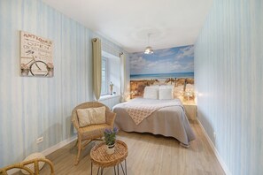 A bedroom with a beach mural, a wicker chair, a bedside table with a plant, and a bed with a patterned blanket. - Domaine de la Dauvinerie – Escapade Cottage (Gratot)