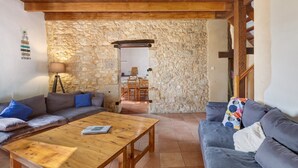 A cozy living room with a stone wall, wooden ceiling beams, a sofa, a coffee table, and a dining area with chairs.