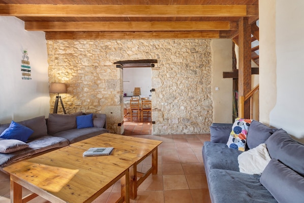 Una sala de estar acogedora con pared de piedra, vigas de madera en el techo, un sofá, una mesa de centro y un comedor con sillas.