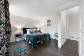 Een slaapkamer met een bed, nachtkastjes, een stoel, een deur en een muur met een afbeelding.