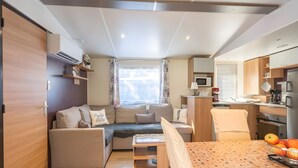 A compact living space with a sofa, dining table, kitchenette, and a window with curtains. - Mobile Home 'Mh324 La Tremblade' with Shared Pool, Private Terrace, and Air Conditioning (La Tremblade)