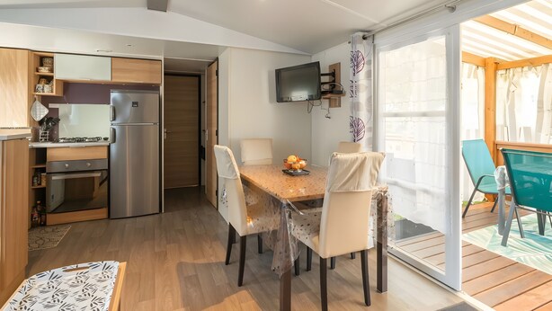 A modern kitchen with a dining area, a TV mounted above the table, and a sliding glass door leading to an outdoor patio. - Mobile Home 'Mh324 La Tremblade' with Shared Pool, Private Terrace, and Air Conditioning (La Tremblade)