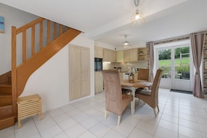 Une cuisine avec des meubles en bois, une table à manger avec des chaises, un escalier et une vue à travers une porte vitrée.