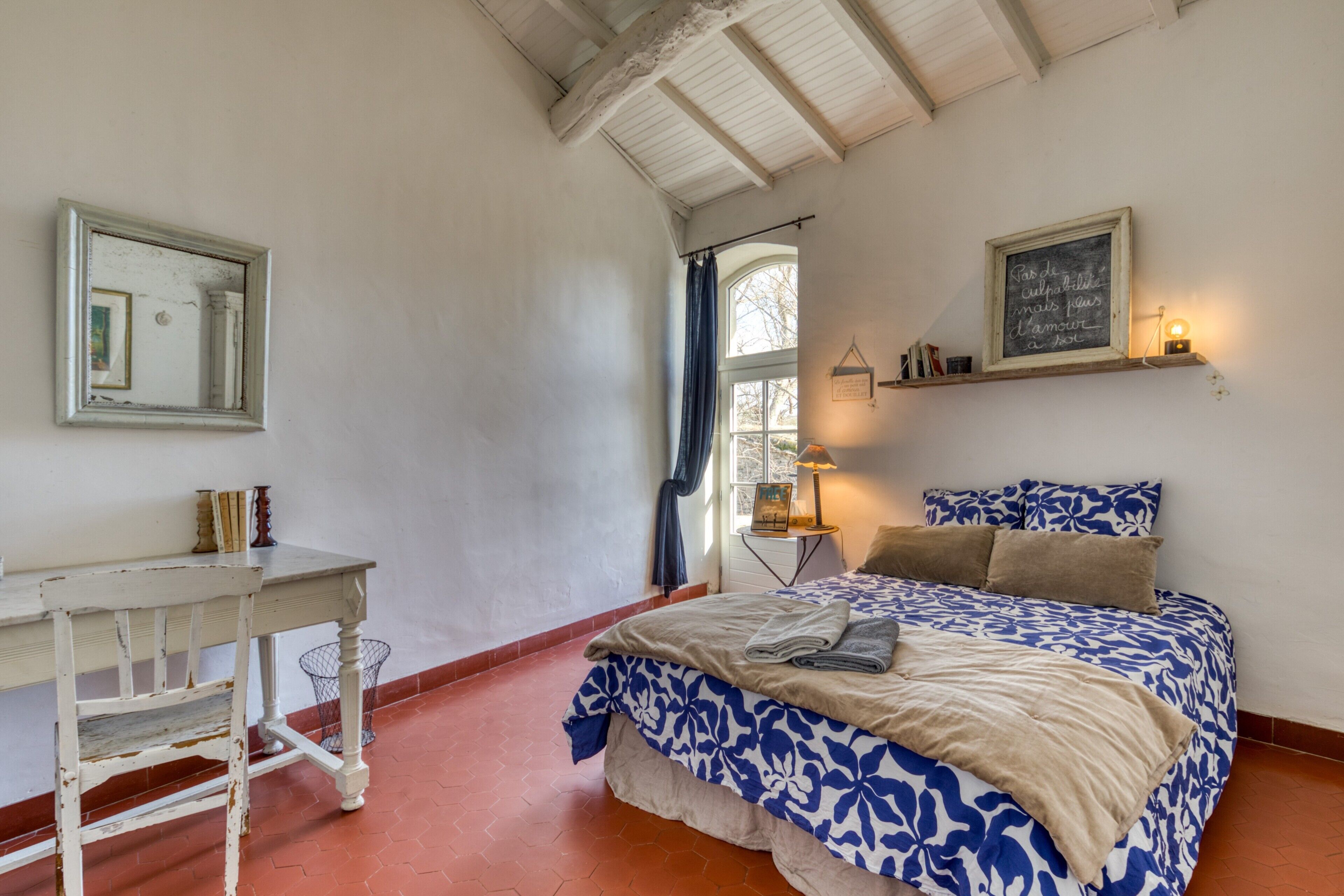 Un dormitorio con cama, escritorio, silla, ventana con cortinas, estante de pared con libros y un cuadro enmarcado.