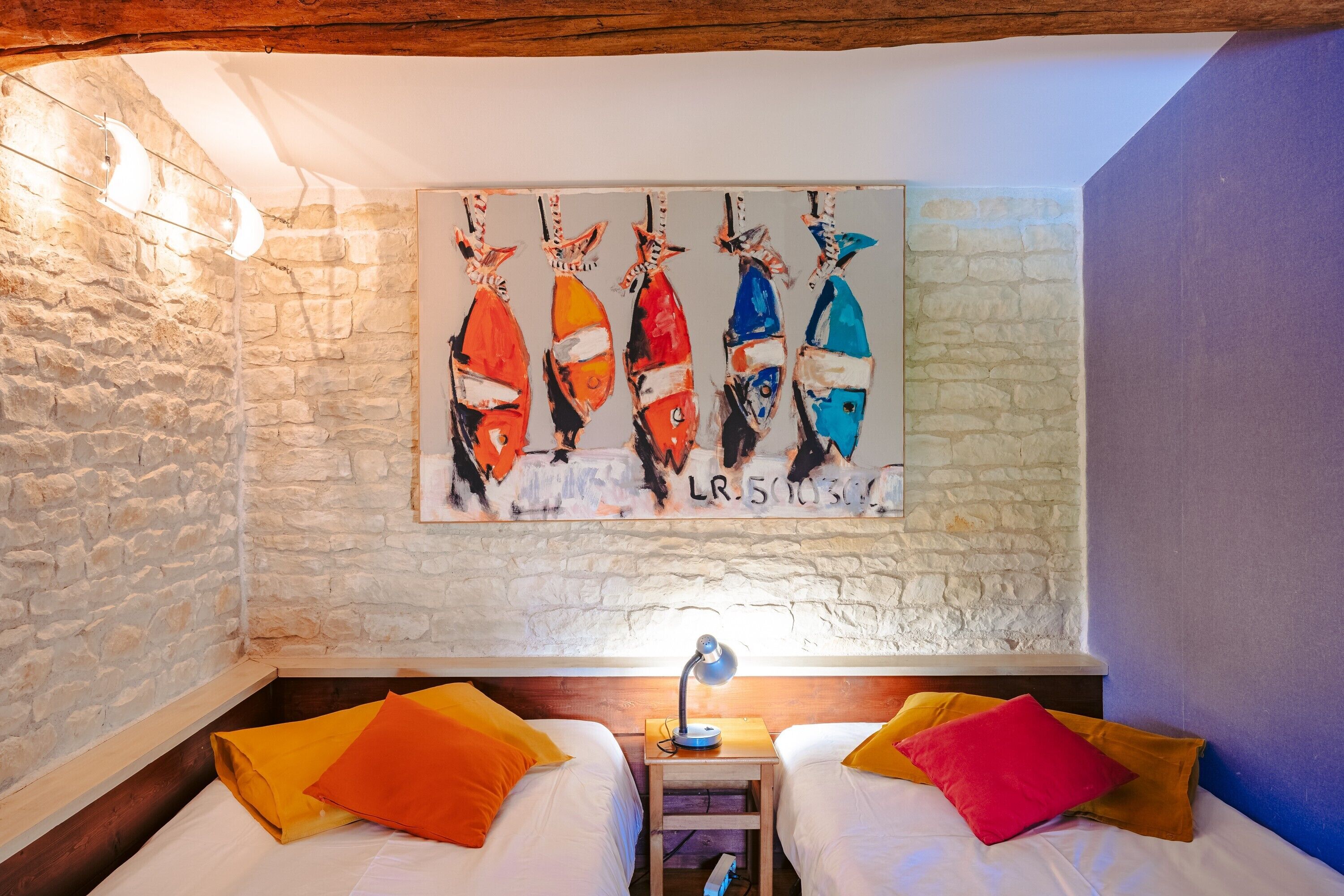 Ein Zimmer mit zwei Betten, einer Steinwand, einem Bild von Fischen und einem kleinen Tisch mit einer Lampe.