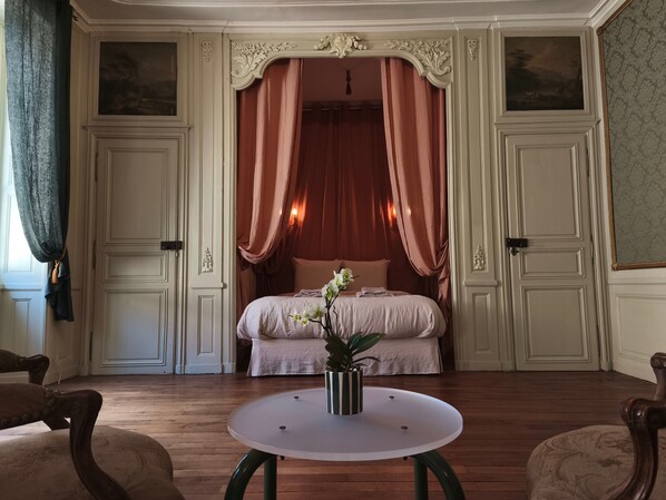Una habitación con cama, cortinas, un florero con flores, una mesa y sillas.