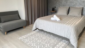 Een moderne slaapkamer met een bed, grijze bank, een tapijt met patroon en een wandplank met speaker.