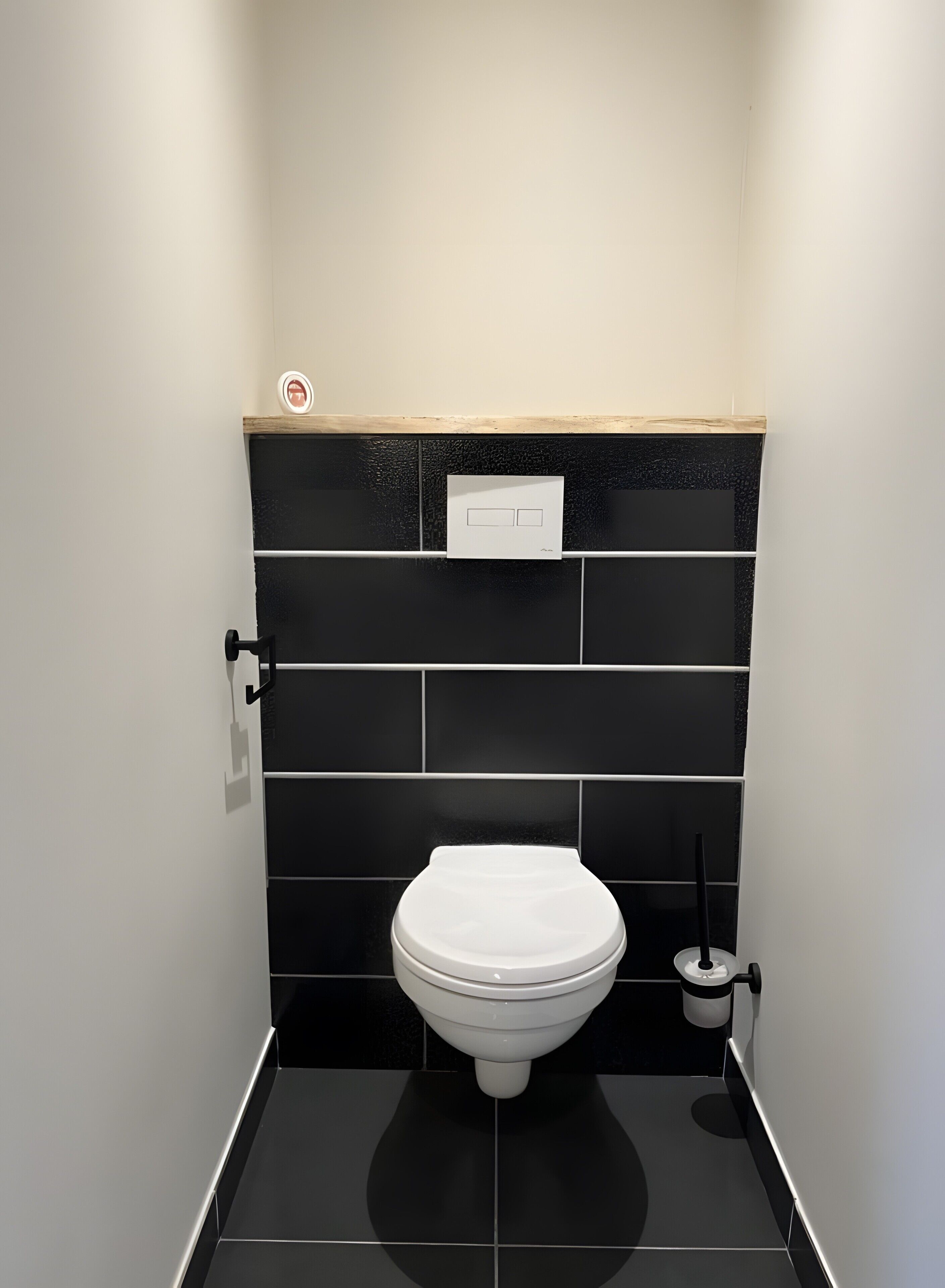 Une salle de bain moderne avec des toilettes blanches, des murs recouverts de carreaux noirs et une étagère en bois au-dessus des toilettes.