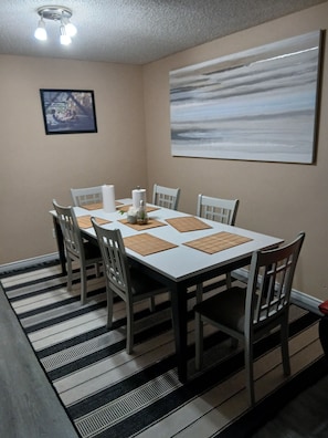 Ein Esszimmer mit weißem Tisch, sechs Stühlen, einem schwarz-weißen Teppich, einem Gemälde an der Wand und einer Deckenleuchte.