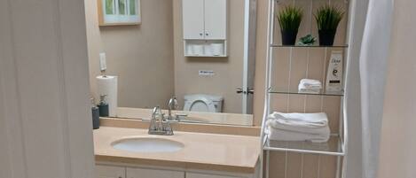 Baño con lavamanos, espejo, inodoro y repisas con artículos de aseo.