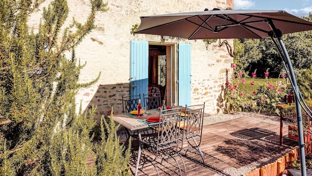 Un edificio rústico de piedra con una mesa y sillas en el patio, una puerta azul y un jardín lleno de flores.