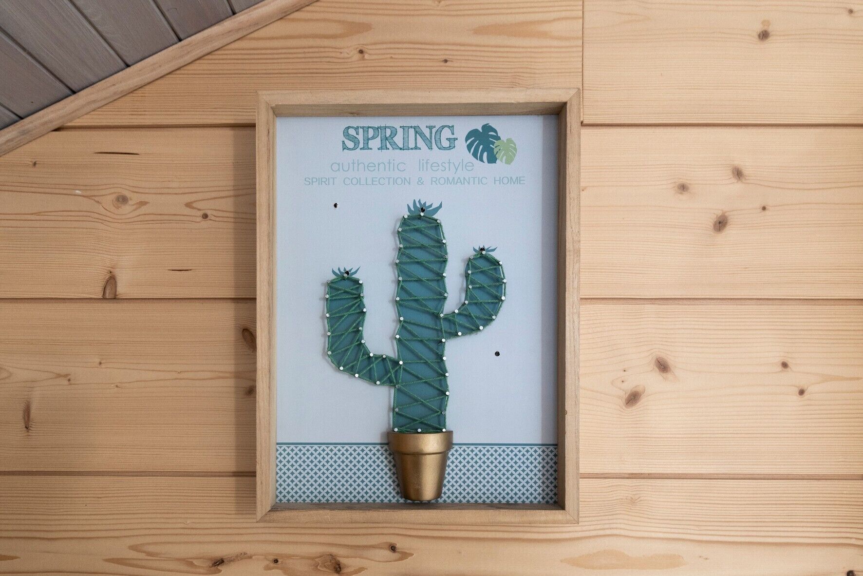 Une œuvre d’art représentant un cactus, encadrée, avec l’inscription « SPRING authentic lifestyle SPIRIT COLLECTION & ROMANTIC HOME » sur un mur en bois.