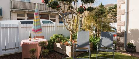 A backyard with a white fence, a tree with red flowers, two metal chairs, a colorful umbrella, and a small table with a bottle.
