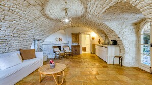 Un interior rústico con techo de piedra, un sofá blanco, sillas de mimbre, una mesa redonda y una zona de cocina con una puerta de madera que indica 'ON'.