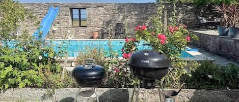 A backyard with a stone wall, a blue slide, a barbecue grill, and a swimming pool.