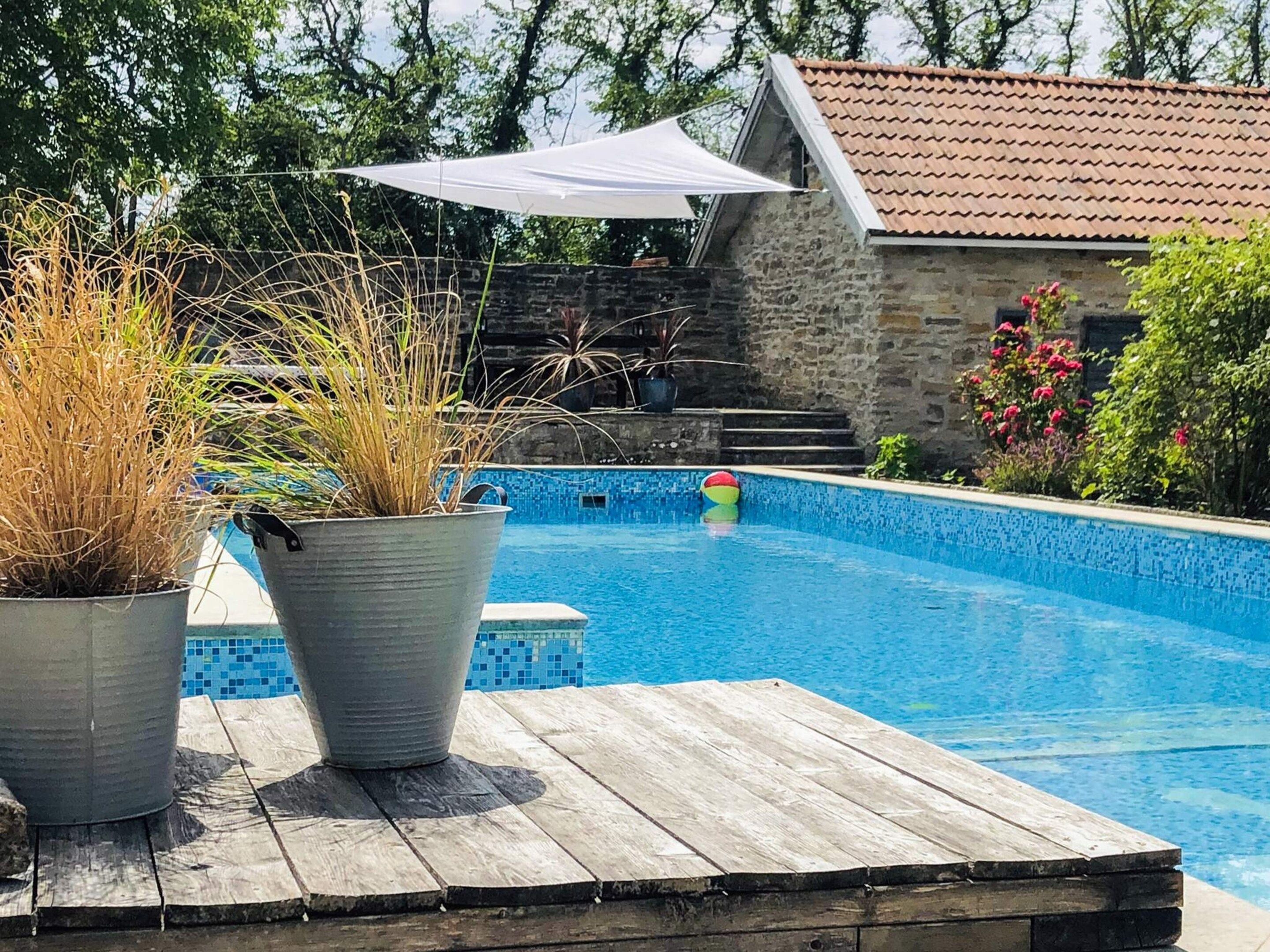 Una piscina amb un pont de fusta, plantes en testos i un edifici de pedra al fons.