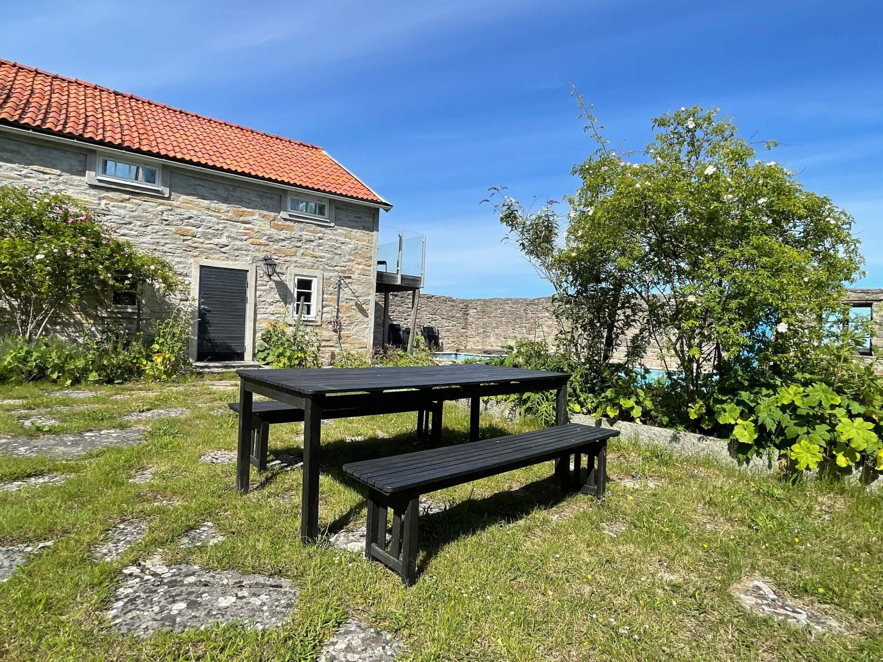 A stone building with a red-tiled roof, a wooden bench, and a grassy area with a tree.