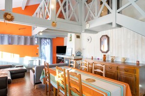 A spacious living area with a high ceiling, a dining table with chairs, a kitchen area, and a television.