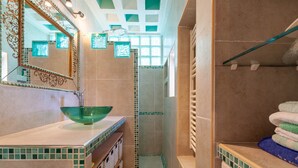 Casa de banho com lavatório de vidro verde, um espelho grande e zona de duche com pavimento em azulejos.