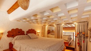 Quarto com cama de madeira, teto com padrão geométrico, janela com cortinas e mesa de cabeceira com abajur.
