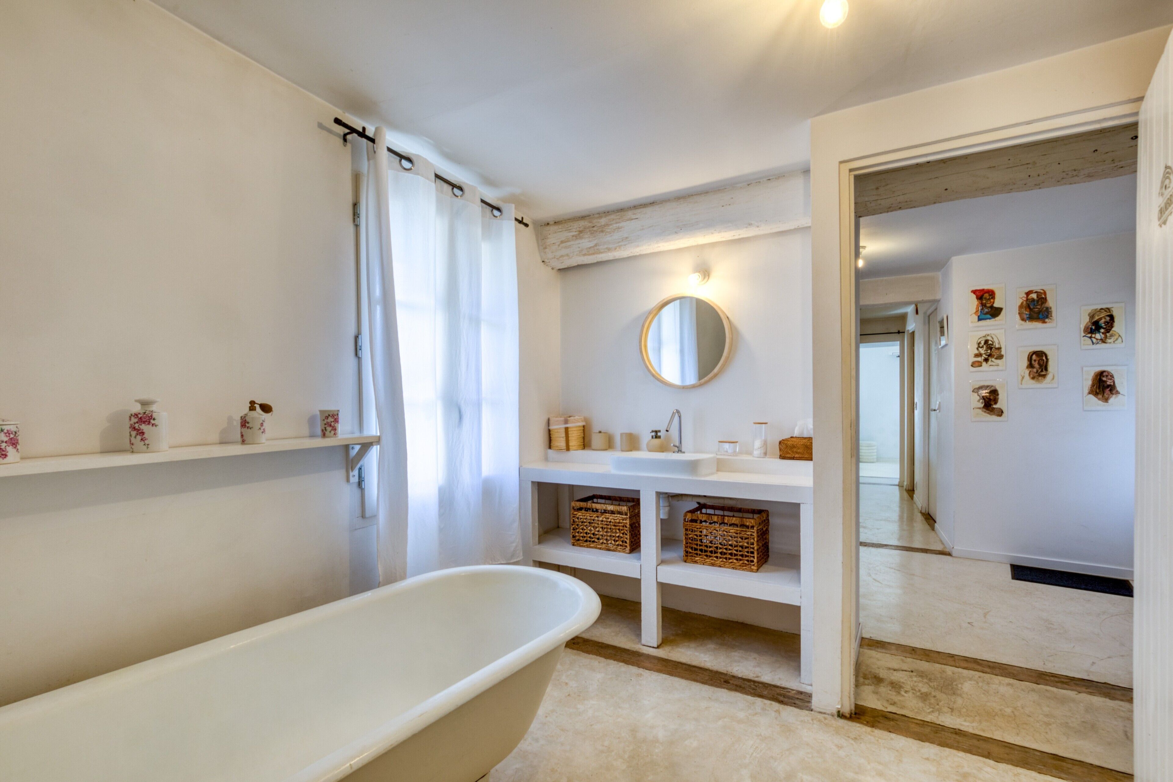 Ein Badezimmer mit einer freistehenden Badewanne, einem Spiegel, Ablagen mit Pflegeprodukten und einer Tür zu einem anderen Raum.