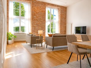 Ein geräumiges Wohnzimmer mit Backsteinwand, großen Fenstern, einer Couch, einem Couchtisch und einem Fernseher.