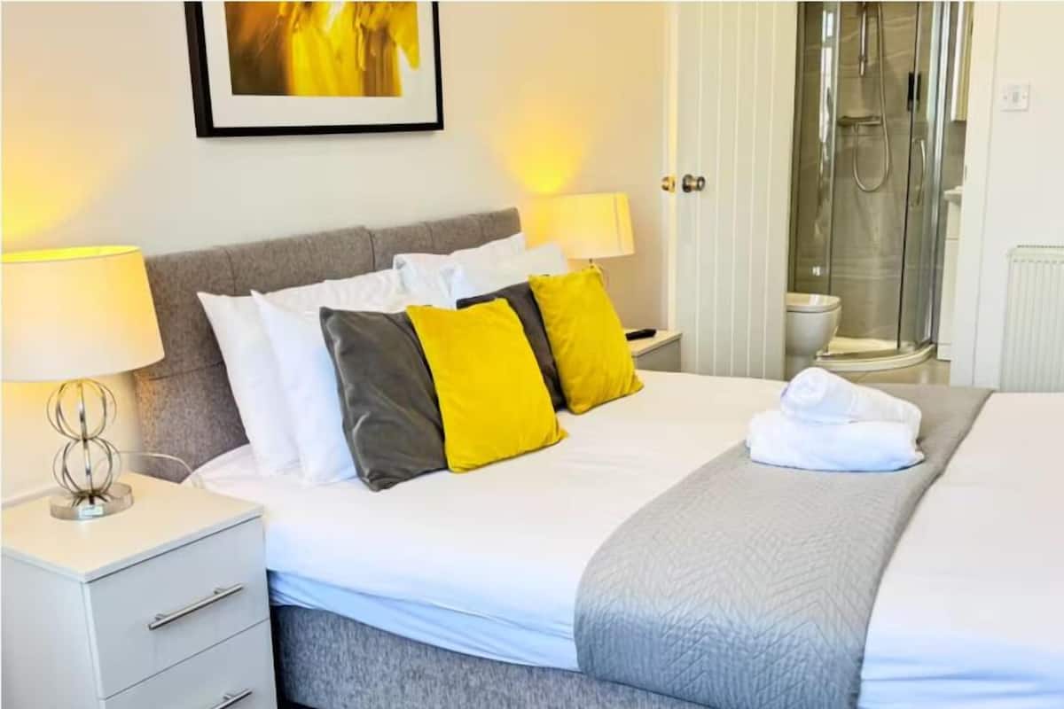 A hotel room with a bed, two bedside lamps, a gray headboard, a white nightstand with drawers, and a framed picture on the wall.