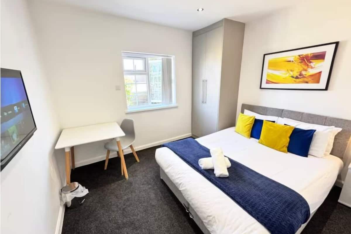 A hotel room with a bed, a desk, a chair, a window, and a painting on the wall.