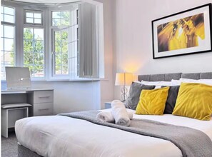 A hotel room with a bed, a desk with a laptop, a window with curtains, and a wall-mounted lamp. - Ox Heathrow Stay  (Hayes)