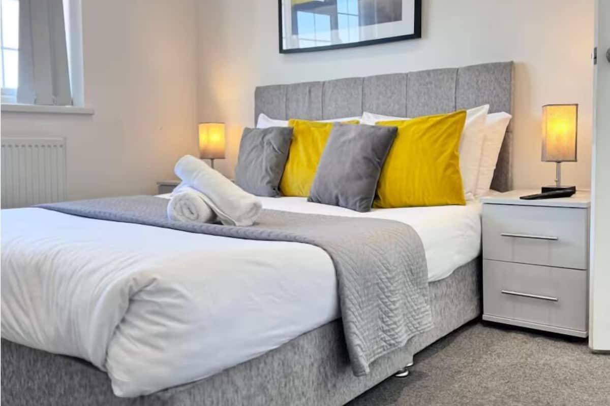 A neatly made bed with a grey headboard, white bedding, and a rolled towel. There are two bedside tables with lamps, a window with curtains, and a framed picture on the wall.