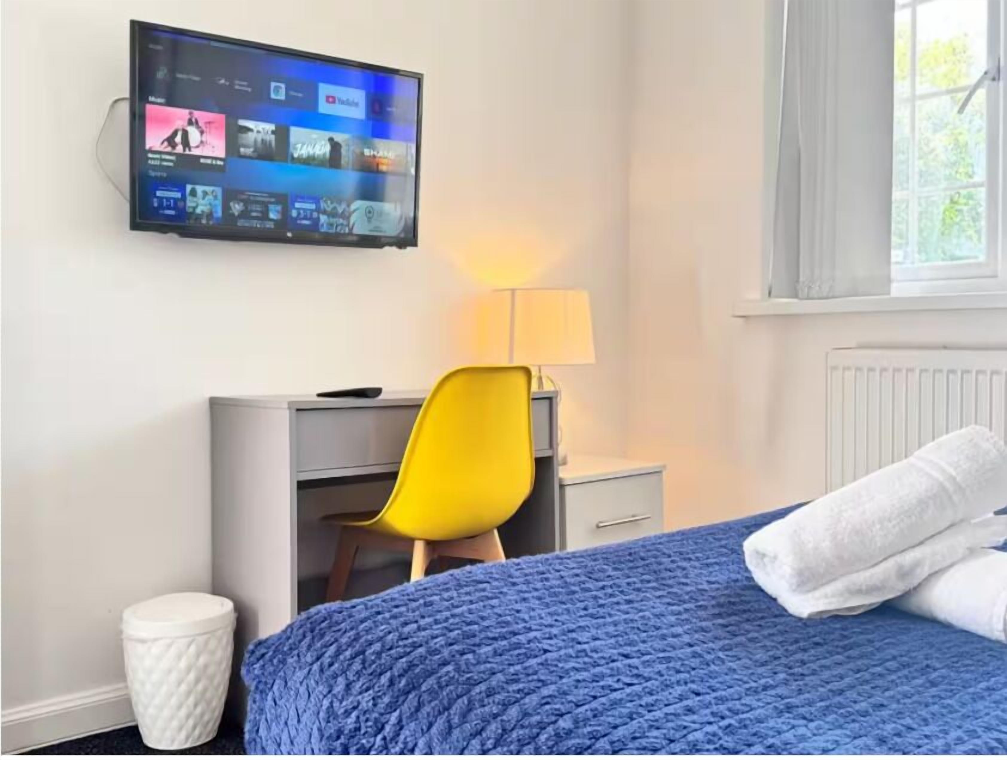 A hotel room with a bed, a desk, a chair, a TV mounted on the wall, and a window with curtains.
