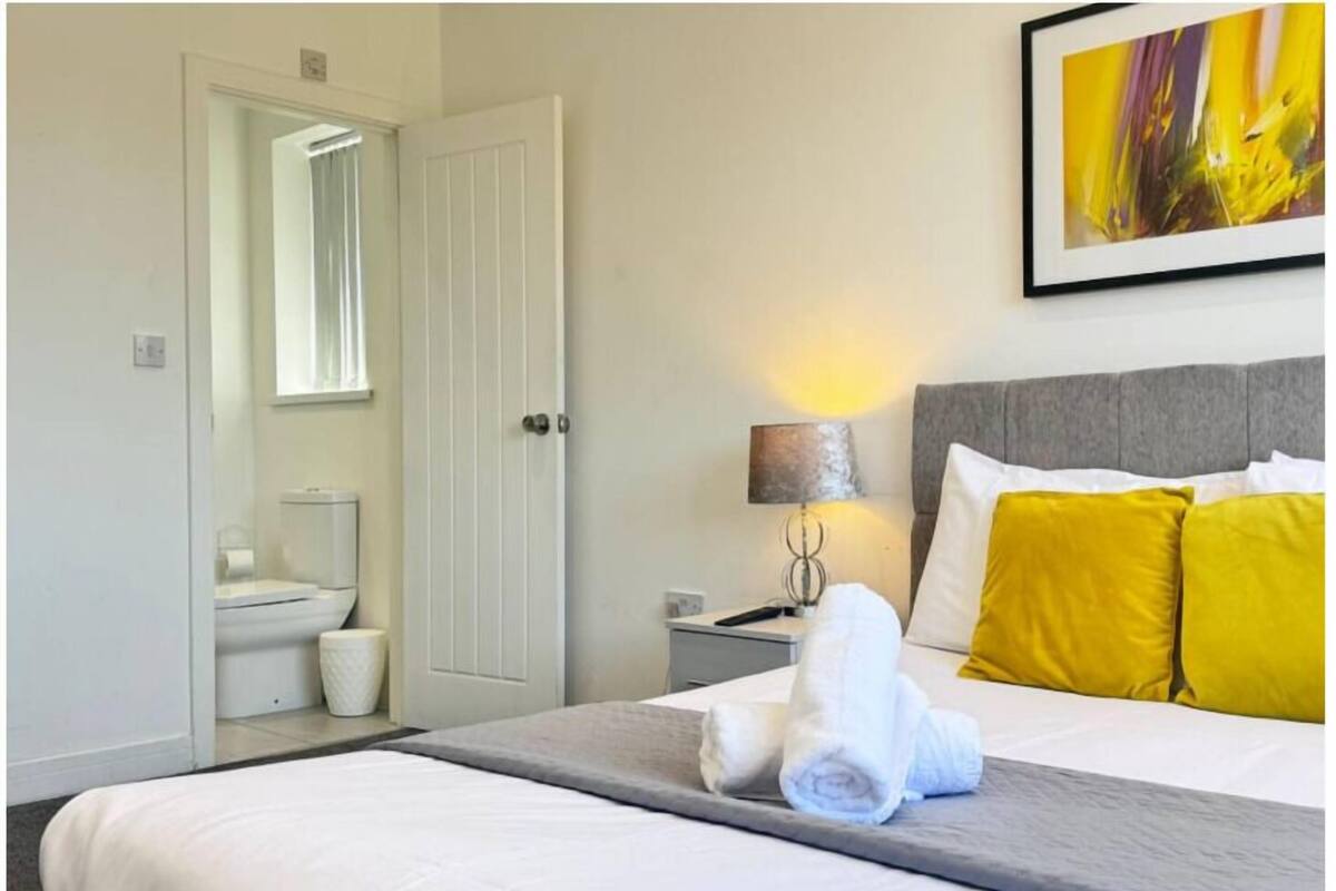 A hotel room with a bed, a nightstand, a lamp, a painting, and a bathroom visible through an open door.