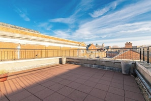 Eine Dachterrasse mit Backsteinmauer, Metallgeländer und blauem Himmel.
