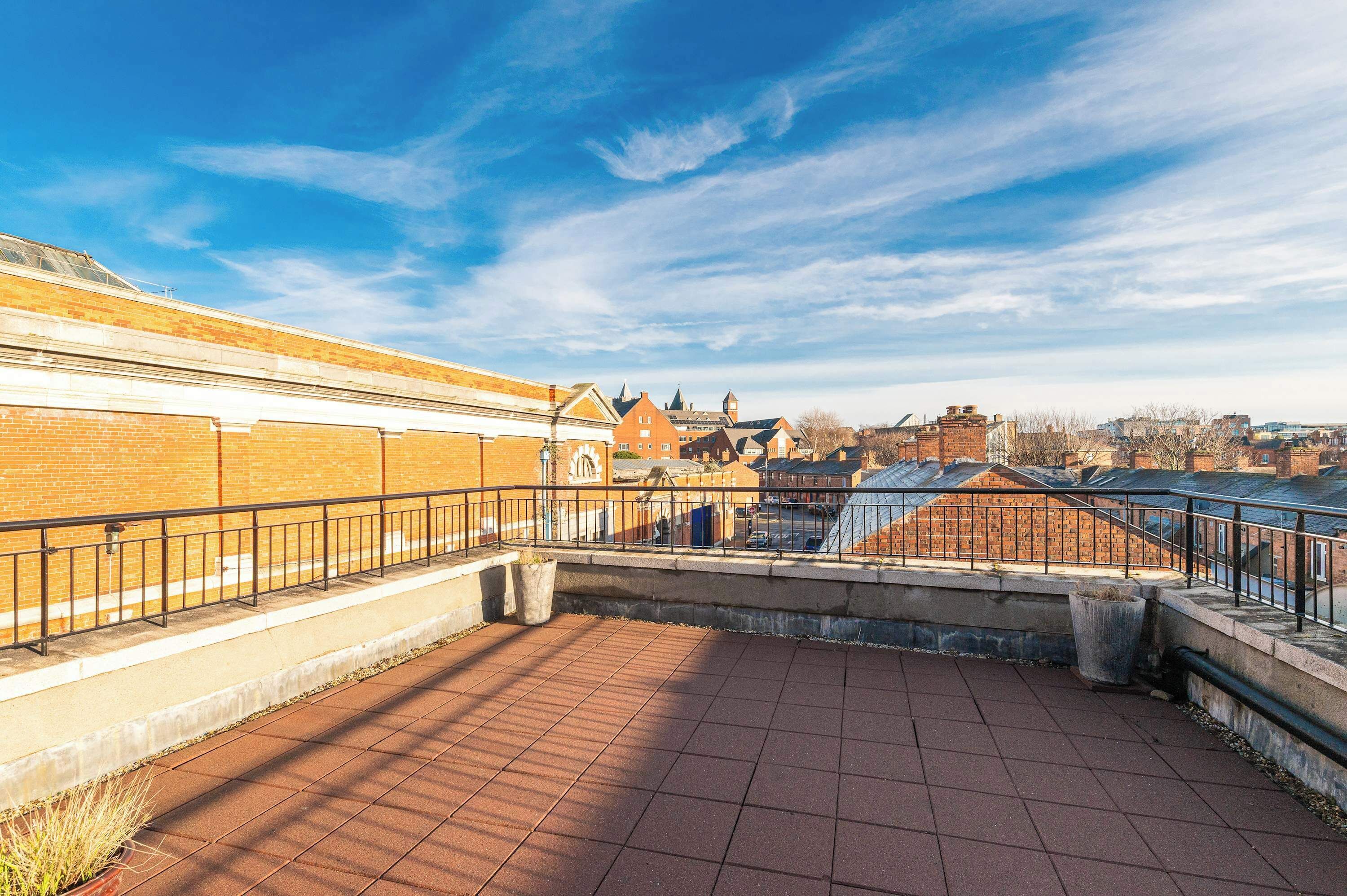 A rooftop terrace with a brick wall, metal railing, and a view of buildings in the background.