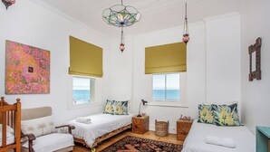 A room with two beds, a chair, a patterned rug, and a large window with a view of the sea.