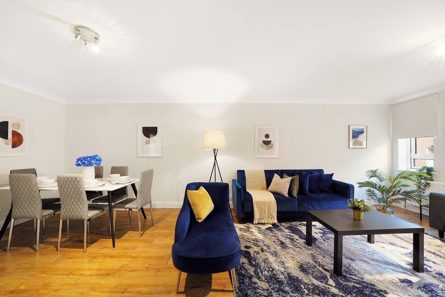 Ein modernes Wohnzimmer mit einem blauen Sofa, einem Esstisch mit Stühlen, einem Couchtisch und einer Stehlampe.