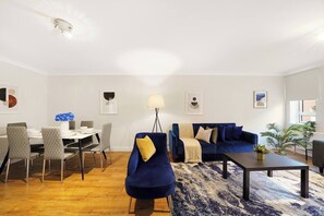 Ein modernes Wohnzimmer mit einem blauen Sofa, einem Esstisch mit Stühlen, einem Couchtisch und einer Stehlampe.