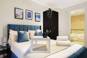 A modern hotel room with a large bed, bedside tables, a small table with a vase, and a bathroom visible through an open door.