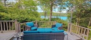 Eine Terrasse mit Korbmöbeln und einem Glastisch, umgeben von Bäumen und mit Blick auf einen See.