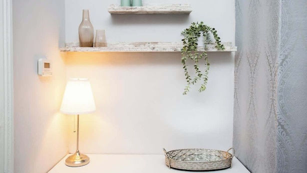Un coin avec un mur blanc, une petite étagère ornée d’objets décoratifs, une plante suspendue, une lampe et un plateau à motifs.