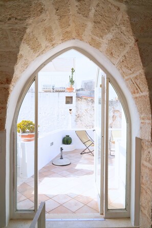 Une porte en arc donnant sur un balcon doté d’un sol carrelé, d’une chaise et de plantes en pot.